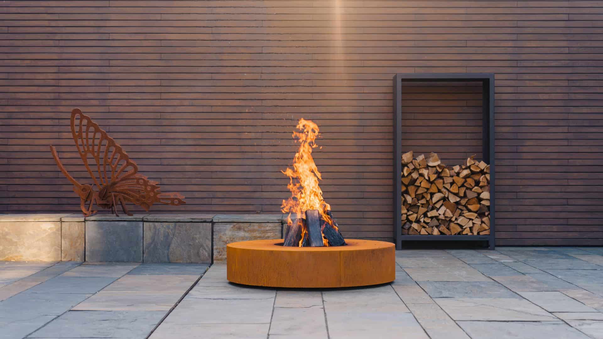 Outdoor fire pit with flames, metal bench, and stacked wood against a wooden wall.