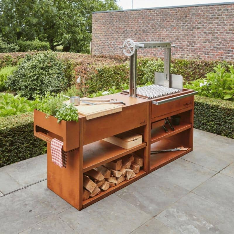 Wooden outdoor kitchen island with grill and storage shelves in a garden setting.