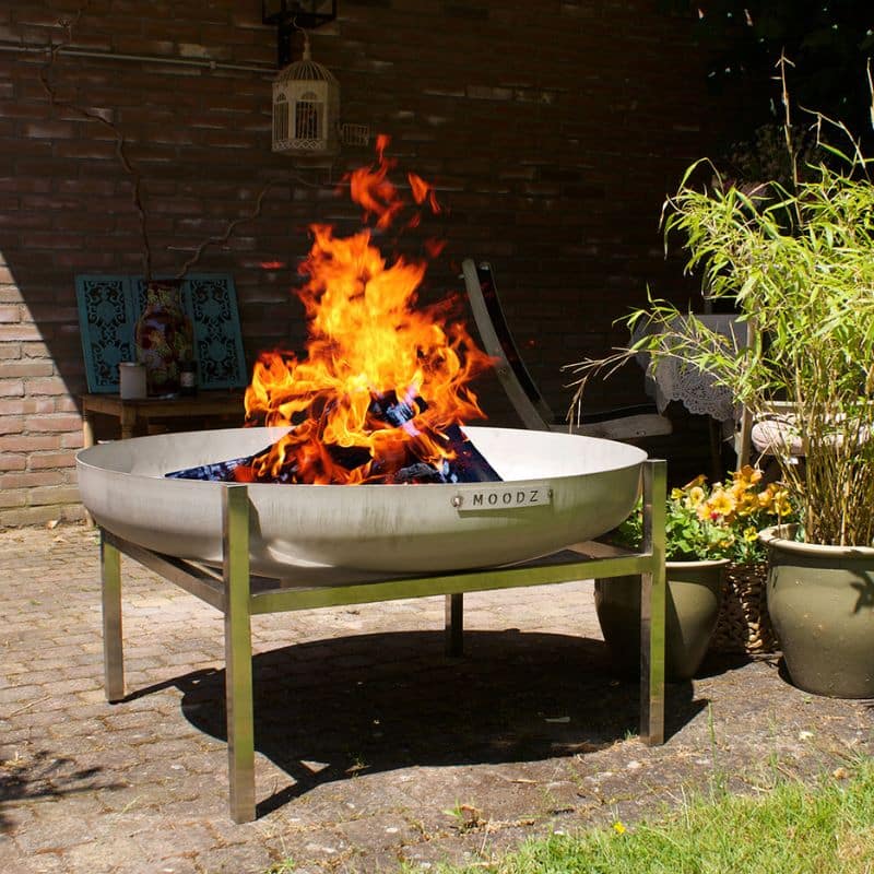 Fire pit with flames in a garden setting with plants and a bench.